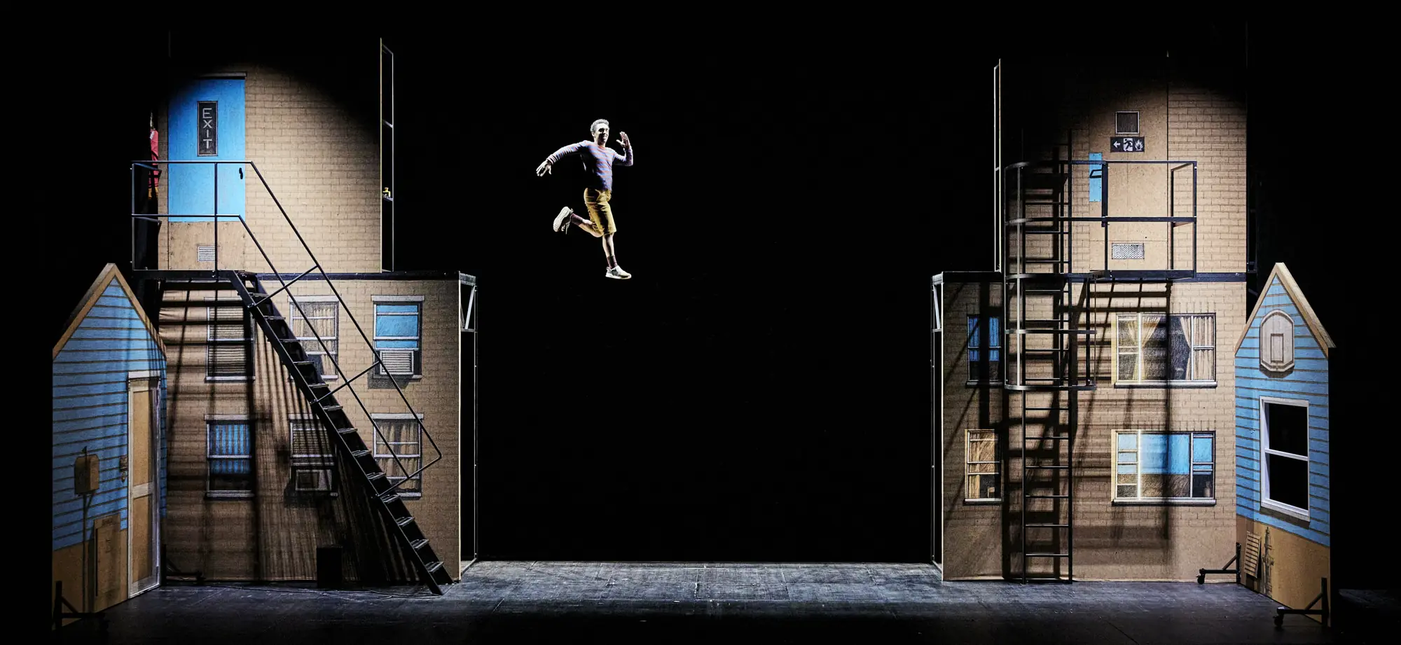 Comédien en lévitation entre deux façades de décor, sur un plateau de théâtre